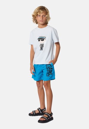 Blond jongetje staat met één hand in de zak, draagt een wit T-shirt met print, felblauwe korte broek met zwarte tekst en zwarte merk-sandalen.