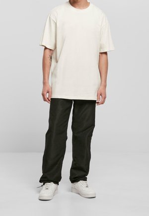 Man draagt een oversized effen beige T-shirt, zwarte rechte broek en witte sneakers, staand tegen een lichtgrijze achtergrond.