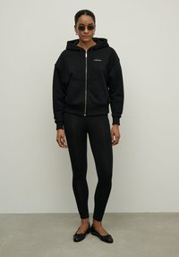 Juodas zip-up džemperis su priekinėmis kišenėmis, derinamas su juodomis leginsomis ir plokščiais batais; akiniai nuo saulės su minimalistiniu dizainu.