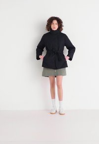 Jachetă neagră cu quilting, cu guler înalt și talie marcată de o curea, purtată peste o fustă verde deschis. Modelul poartă șosete și pantofi albi, stând în fața unui perete alb.