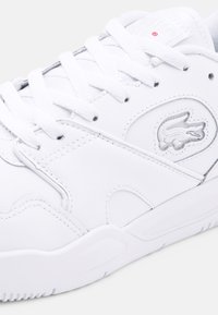 Valged sneakers, mis on valmistatud siledast nahast, lamepaeltega ja tekstureeritud kummist tallaga. Includes hall Lacoste logo ja õmblusdetailid.