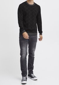 Zwarte sweater met diamanten patroon, ronde halslijn en ribgebreide manchetten, gecombineerd met grijze jeans en zwarte sneakers met witte zolen.