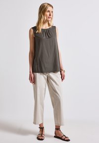 Modellen poserar i en ärmlös mörk topp och ljusa byxor, iförd sandaler med remmar. Den mjuka bakgrunden framhäver en avslappnad, stilren look.
