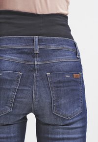 Jeans de denim elástico azul-escuro com uma cintura alta preta e costuras contrastantes. Apresenta dois bolsos traseiros e uma pequena etiqueta de logótipo.