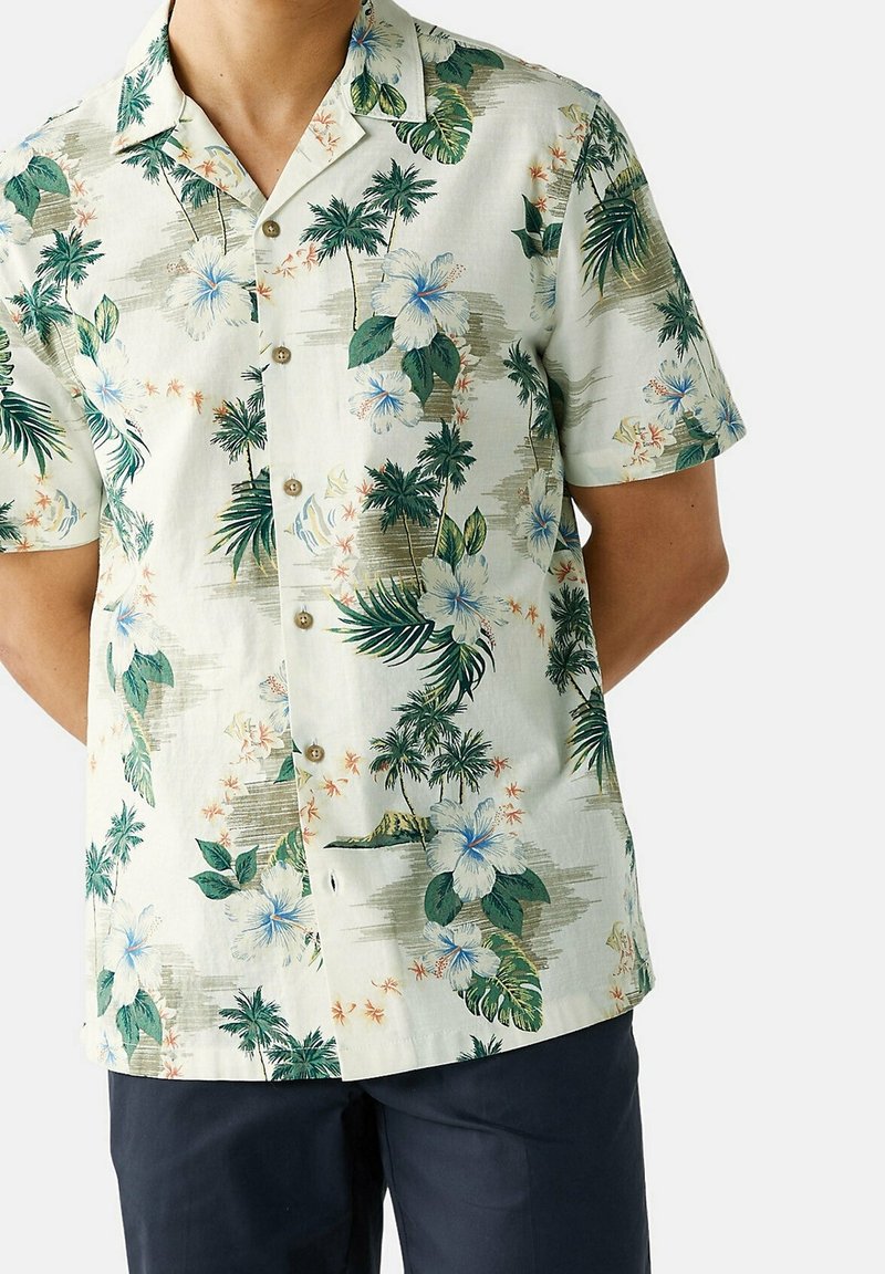 Homme portant une chemise blanche à manches courtes à boutons avec un imprimé de palmiers verts et de fleurs d'hibiscus bleues, associée à un pantalon foncé.