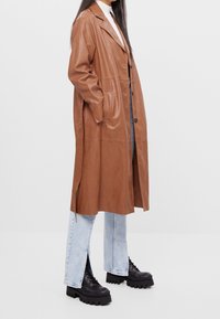 Hnědý kožený trenčkot s přizpůsobeným střihem, bočními kapsami a zapínáním na knoflíky. V kombinaci se světle modrými džínami se spadlým lemem a černými masivními botami.