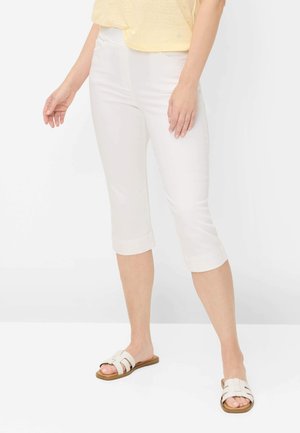 Kvinde iført hvide korte bukser, en lyst gul top og hvide slip-on sandaler, stående mod en ensartet lys baggrund.