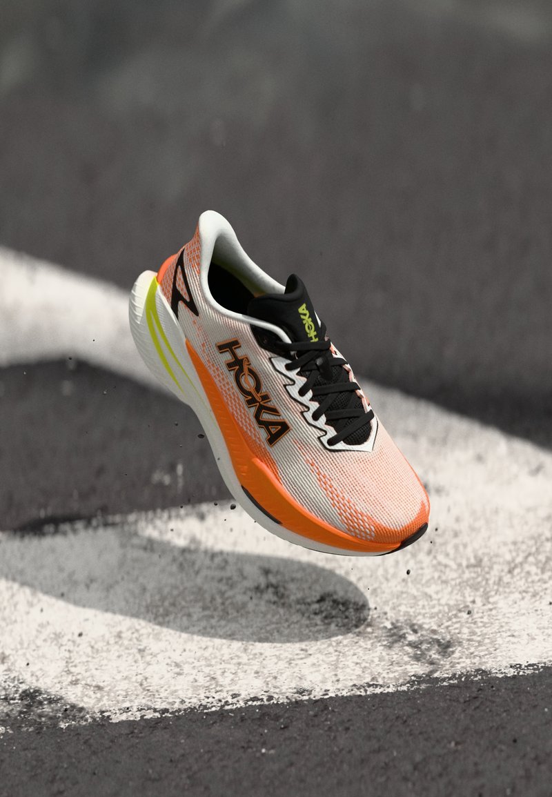 Sapato de corrida Hoka em branco e laranja com atacadores e sola pretos, pairando sobre uma superfície de asfalto com sinalização rodoviária branca.
