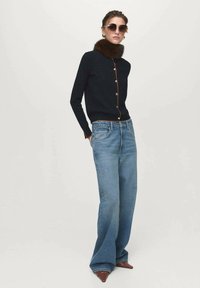 Cardigan navy con colletto in pelliccia sintetica, bottoni dorati, abbinato a jeans a gamba larga azzurri e décolleté a punta marroni. Occhiali da sole e orecchini inclusi.
