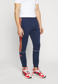 Námořnické modré sportovní kalhoty s oranžovými pruhy po stranách, bílým lemem na manžetách a vyšitým logem na levé noze.