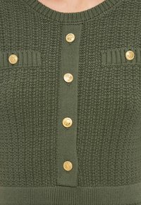 Ζακέτα από πλεκτό σε ελιά πράσινο με υφή, χρυσά κουμπιά και οριζόντιες ριγωτές λεπτομέρειες κοντά στις τσέπες στο στήθος.