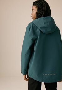 Veste imperméable teal avec capuche, offrant une coupe décontractée, des poignets élastiques et un détail en bande réfléchissante au dos.