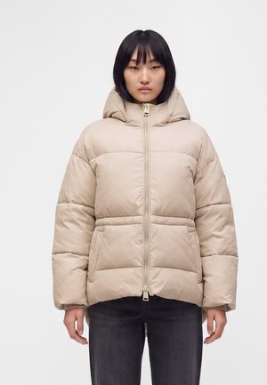 Jeune personne portant une doudoune à capuche beige avec poches avant et fermeture éclair, associée à un pantalon foncé, debout devant un fond uni.