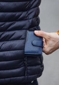 Navy blue leather wallet with a smooth texture, featuring a zipper and a snap closure. Logo embossed on the front. Hand is reaching into a jacket pocket.