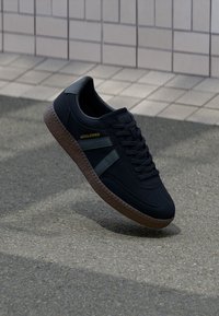 Zapatillas negras con un diseño texturizado, que cuentan con una suela de goma, rayas verde oscuro y detalle del logotipo dorado en el lateral.