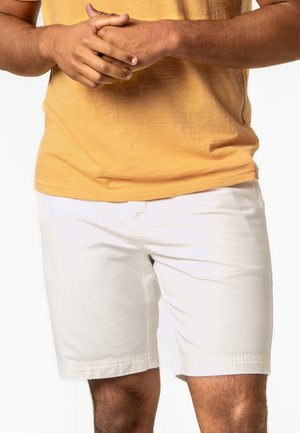 Mann trägt ein gelbes T-Shirt und weiße Shorts, steht mit verschränkten Händen vor der Taille vor einem einfarbigen Hintergrund.