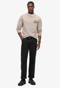 Beige sweatshirt med rund halsudskæring og patchdetalje på venstre bryst, parret med sorte bukser. Modellens på sorte sko.
