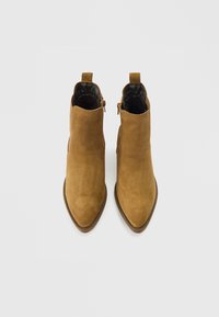 Botas de cano curto em camurça castanha com bicos pontiagudos, dois fechos laterais e pequenas abas de puxar na parte traseira. Textura lisa sem padrões visíveis.