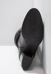 Botte en cuir noire avec un bout rond et un talon angulé, présentant une semelle en caoutchouc texturée avec un motif de feuille et une étiquette de taille embossée.