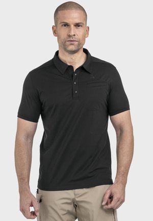 Mann trägt ein schwarzes kurzärmeliges Poloshirt mit Knöpfen und beige Hose, steht vor einem schlichten hellen Hintergrund.