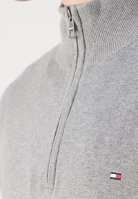 Grauer Reißverschluss-Pullover mit geripptem Kragen, ausgestattet mit einem kleinen gestickten Logo in Rot, Weiß und Blau auf der Brust. Weicher Strickstoff.