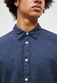 Camisa de botones azul marino hecha de tejido texturizado. Presenta un collar clásico, cinco botones blancos y un sutil detalle bordado.