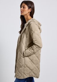 Beige gewatteerde jas met een capuchon, voorzien van rits- en drukknoopsluitingen, zijzakken en een tekstlabel op de mouw. Glad textuur.