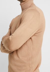 Beige rullekrave sweater med ribbet krave og strukturerede sidelæg med smalle lodrette striber. Fremstillet af et blødt strikstof.