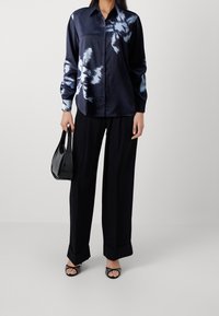 Chemise en satin bleu marine à motif floral bleu clair, boutonnée sur le devant, manches longues, associée à un pantalon noir à jambes larges et un sac à main noir.