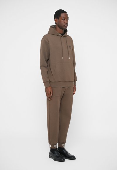 Brązowa bluza z kapturem, posiadająca sznurki i kieszeń z przodu, zestawiona z dopasowanymi spodniami z mankietami i napisami, noszona z czarnymi butami.