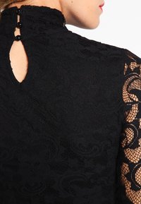 Top de encaje negro con un cuello alto, que presenta un detalle de ojal en la parte posterior y tres botones decorativos negros. Patrón floral intrincado.