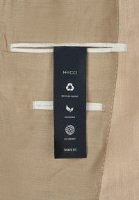 Étiquette écologique sur un tissu beige texturé avec des coutures blanches. L'étiquette présente des icônes pour le contenu recyclé, la durabilité et l'ajustement des formes.