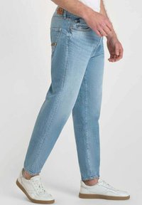 Pantalones vaqueros de denim azul claro con corte recto, cinco bolsillos y parche de cuero marrón en la cintura. Llevados con zapatillas blancas.