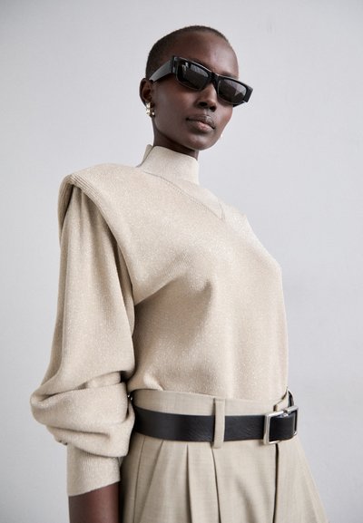 Beige strikbluse med høj hals, pufærmer og subtil glimmer, parret med plisserede beige bukser og et sort læderbælte. Sorte solbriller og øreringe.