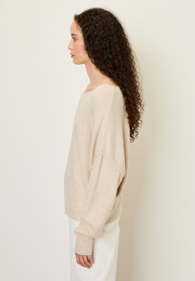 Femme aux longs cheveux bouclés portant un pull beige ample et un pantalon blanc, debout de profil sur un fond clair uni.