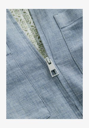 Šviesiai mėlynas tekstūruotas audinys su zigzago raštu, sidabrinis užtrauktuko detalės, o viduje matoma gėlėta pamušalo tekstūra.