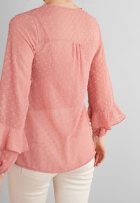 Blouse rose transparente avec une texture à pois, manches évasées, encolure arrondie et un plis dans le dos. Tissu léger.