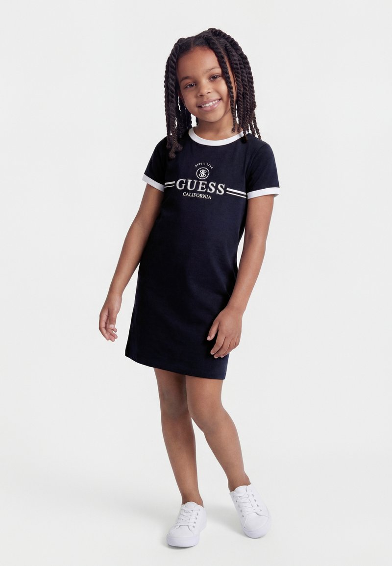 Κορίτσι με πλεγμένα μαλλιά που φοράει μαύρο φόρεμα Guess California με λευκή διακόσμηση και λευκά αθλητικά παπούτσια, στέκεται και χαμογελά σε λευκό φόντο.