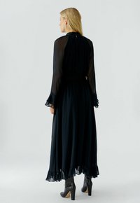Frau mit langen blonden Haaren in einem langen schwarzen transparenten Kleid und schwarzen High-Heels, die seitlich vor einem schlichten weißen Hintergrund steht.