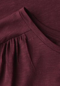 Tela de camiseta burdeos con una textura suave, que presenta un escote en forma de barco y detalles fruncidos a lo largo de la costura del hombro.