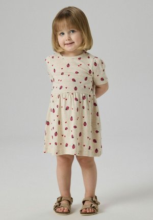 Junges Mädchen mit blondem Bobhaarschnitt, das ein beiges Kleid mit Marienkäfern und Herzmotiven trägt, steht mit den Händen hinter dem Rücken und lächelt.