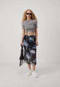 Sivi crop top s nabrancima i kratkim rukavima, u kombinaciji s crnom prozirnom maksi suknjom s cvjetovima. Model nosi bijele tenisice i drži crnu torbu.