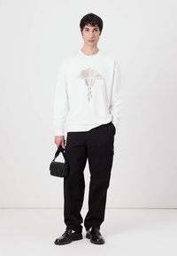 Sudadera blanca con un diseño geométrico beige, pantalones cargo negros y zapatos negros. Sosteniendo un pequeño bolso negro. Fondo simple.