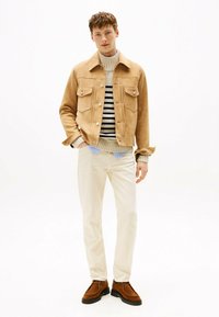Chaqueta de ante marrón con dos bolsillos frontales, suéter de punto a rayas, pantalones crema y zapatos marrones, de pie contra un fondo blanco.