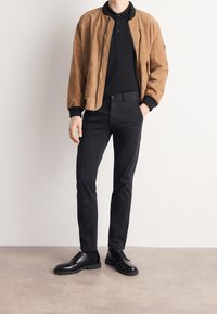 Brown suede bomber jacket over a black polo shirt, paired with black slim-fit pants and black leather shoes with a thick sole.