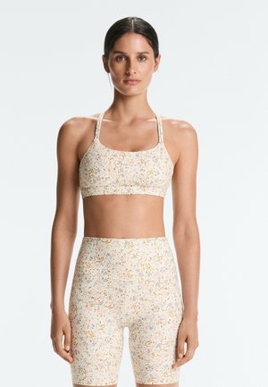 Femme aux cheveux foncés attachés en arrière, portant un soutien-gorge de sport assorti à motif floral pâle et un short de vélo taille haute, debout sur un fond blanc.