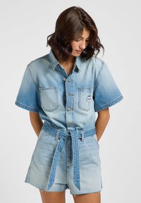 Ljusblå denimskjorta med korta ärmar, två bröstfickor och knäppning framtill. Kombinerat med högmidjade denimshorts och ett knytbälte i midjan.