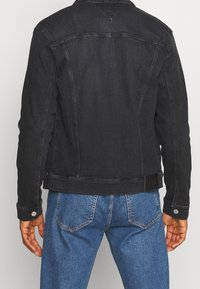 Person som bär en svart jeansjacka och blå jeans, sedd bakifrån mot en ensam ljus bakgrund.