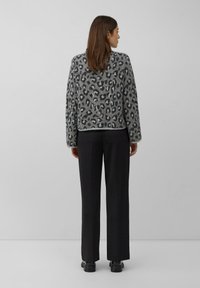 Vrouw met bruin haar kijkt weg, gekleed in een grijze leopardprint trui, zwarte rechte broek en zwarte schoenen tegen een effen grijze achtergrond.