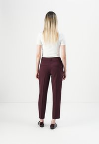 Žena s dlouhými blond vlasy, oblečená v bílém tričku s krátkým rukávem, tmavě burgundských kalhotách na míru a tmavých botách, stojící zády.